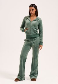 Der Velour-Trainingsanzug in gedecktem Grün verfügt über eine Zip-Hoodie und weit geschnittene Hosen mit Fronttaschen und Kordelzug, hat eine weiche Textur und eine taillierte Passform.