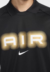 Svart polo med en stor stiliserad "AIR"-grafik i guld och vitt, samt en liten vit Nike-logotyp på den nedre delen.