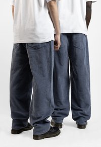 Pantaloni in corda a gamba larga di colore blu navy con una texture a righe. Presentano due tasche posteriori e sono indossati con scarpe nere.