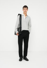 Jonge man in een lichtgrijze polo met lange mouwen, zwarte broek, zwarte schoenen, met een zwarte leren schoudertas op een witte achtergrond.
