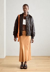 Schwarze Bomberjacke, weißes Croptop, eleganter orangefarbener Gürteltasche, beiger, fließender Rock und klobige schwarze Sandalen, vor neutralem Hintergrund.