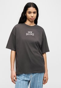 Γυναίκα με μακριά σκούρα μαλλιά, φορώντας μια φαρδιά σκουρόγκριζα μπλούζα New Balance και ανοιχτό μπλε τζιν με ρίγες, μπροστά σε απλό φόντο.