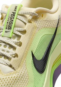 Close-up van een beige en lichtgroene Nike Pegasus Premium sneaker met een zwarte swoosh en patroonveters door textuurrijke veterogen.