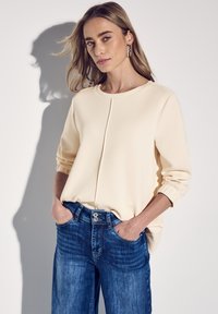 Cremfarbener Pullover mit rundem Halsausschnitt, langen Ärmeln und einer Mittelnaht. Kombiniert mit blauen Jeans aus Denim mit metallischem Knopfverschluss.