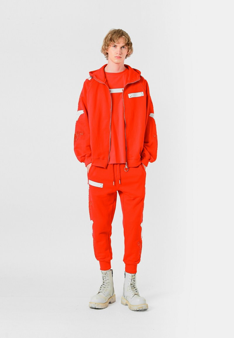 Conjunto de sudadera roja con capucha y pantalones deportivos con detalles bordados y acentos en blanco, combinado con botas gruesas de color claro. Textura suave.