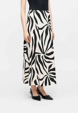 Frau trägt ein schwarzes ärmelloses Oberteil, einen langen weißen Rock mit schwarzem abstraktem Blättermuster und schwarze spitze Slingback-Ballerinas mit Schleifen.