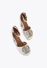 Sandali beige con tacco, con tomaia texture, punta aperta, cinturino alla caviglia e una fibbia decorativa impreziosita da strass sul davanti.