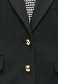 Mustassa bleiserissä on kiiltävät kultakolikot, jämäkät läpät ja houndstooth-kuvio alla. Siinä on kaksi etutaskua ja sileä kangaspinta.