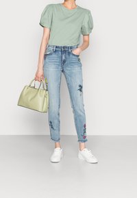 Mujer vistiendo una blusa de manga abullonada verde claro, jeans bordados con flores en azul claro, zapatillas blancas, sosteniendo un bolso verde claro, de pie sobre un suelo gris.