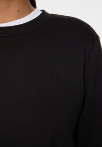 Sweatshirt noir avec un col rond côtelé et un logo en relief subtil sur la poitrine. Le tissu semble lisse et est légèrement texturé.