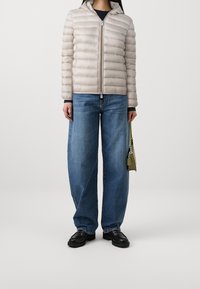 Hellbeige Pufferjacke mit Reißverschluss und gerippten Bündchen, kombiniert mit weit geschnittenen blauen Jeans und schwarzen Schuhen, während sie eine grüne Handtasche hält.