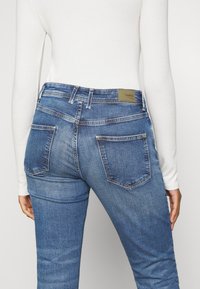 Blå denimjeans med medelhög midja, som har två bakfickor och en läderlapp på midjebandet. Ytan är slät, färgen är jämn.