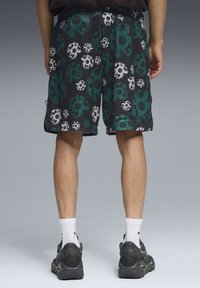 Shorts de malla negros con un patrón de calaveras y flores en teal y blanco. Cintura elástica, bolsillos laterales y ribete negro. Estilizados con zapatos grises.
