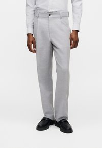 Άνδρας που φοράει ανοιχτό γκρι παντελόνι φόρμας, λευκό πουκάμισο με κουμπιά και μαύρα μοκασίνια, στέκεται μπροστά σε απλό λευκό φόντο.