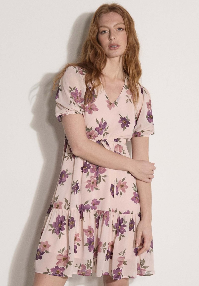 Femme rousse portant une robe rose clair à imprimé floral violet, manches courtes bouffantes et col en V, debout contre un mur uni blanc cassé.