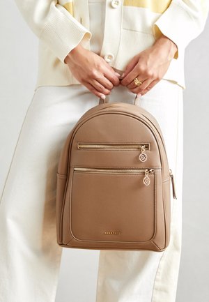 Handen houden een beige leren rugzak met gouden ritsen en logo, gedragen met crèmekleurige broek en een crème-gele gestreepte trui.