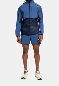 Personne portant une veste zippée bicolore bleue et un short assorti avec des chaussures de course beiges, se tenant devant un fond clair.