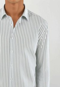 Mann iført en hvit skjorte med tynne svarte vertikale striper, med åpen krage, som står mot en enkel lys bakgrunn.