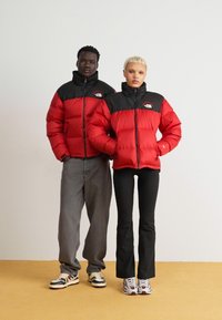 Røde puffjacketer med sorte skuldre, lynlåslukning og logodetaljer, parret med grå jeans og sorte flared bukser. Farverige sneakers er iført.