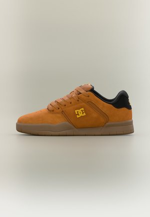 Baskets basses en daim marron avec semelle en gomme, col rembourré noir, lacets assortis et logo DC jaune sur le côté et le talon.