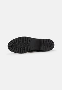 Suela de zapato de goma negra con un patrón de relieve profundo, forma ligeramente curvada y superficie texturizada. Presenta un detalle de logo y bordes redondeados.