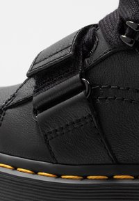 Černá kožená bota s suchým zipem, texturovaným svrškem, kontrastním žlutým prošíváním a odolnou podrážkou. Zaměření na design a funkčnost.