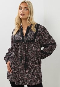 Blusa nera con motivi floreali in rosa e bianco, con uno yoke nero in tessuto testurizzato, lacci con pon pon e lunghe maniche a sbuffo.