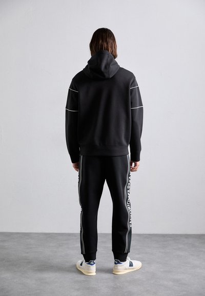 Sweatshirt preta com costuras brancas, combinada com calças de moletão pretas com marcação lateral. O modelo usa ténis creme e azul-marinho.