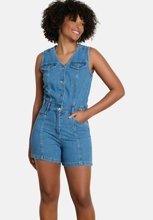 Femme aux cheveux bouclés souriante, portant une combinaison bleu denim sans manches boutonnée avec des poches, debout avec une main dans une poche.