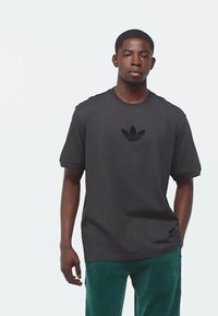 Černé bavlněné tričko s kulatým výstřihem a krátkými rukávy, s tón v tónu logem Adidas na prsou. Nosí se s zelenými kalhotami.