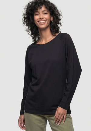 Smiling woman with curly hair wears black long-sleeve shirt and olive green pants, standing against a plain light background.