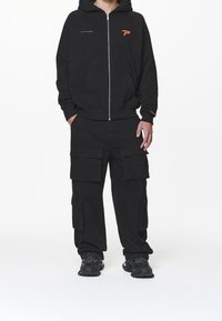 Felpa nera con zip e logo arancione, abbinata a pantaloni cargo neri dotati di molteplici tasche. Footwear comprende sneakers nere.