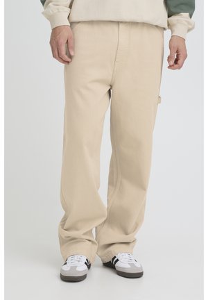 Mand iført beige løstsiddende bukser og hvide sneakers, stående på en ensartet lys baggrund.