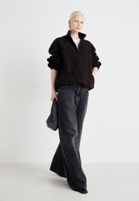 Schwarze Fleecejacke mit hohem Kragen, Reißverschluss, oversized Passform, kombiniert mit grauen weit geschnittenen Jeans und einer strukturierten Clutch-Handtasche.