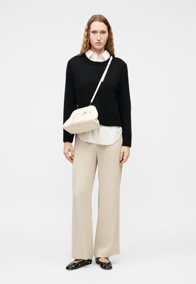 Γυναίκα με μακριά κυματιστά μαλλιά φοράει μαύρο πουλόβερ πάνω από λευκό πουκάμισο, μπεζ παντελόνι με φαρδύ πόδι, μαύρες μπαλαρίνες με τρουκς και κρεμ τσαντάκι ώμου.