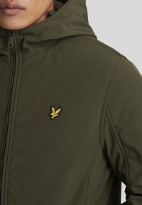 Olivegrønn hettegenser laget av glatt stoff, med glidelås og en liten gul brodert logo på venstre bryst.