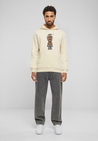 Krem hettegenser med et grafisk trykk av en tegneseriefigur i midten, kombinert med mørkegrå jeans og hvite sneakers, i et studio.