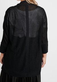 Černý, průhledný, nadměrný kardigan s měkkou texturou, dlouhými rukávy a rovním lemem. Obsahuje zadní šev běžící od shora dolů.