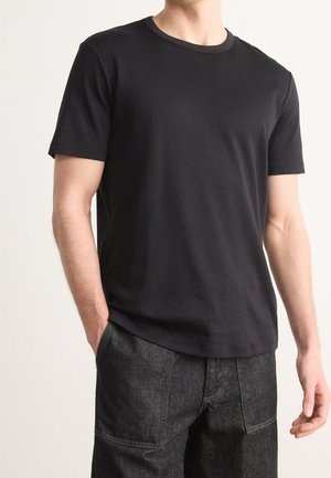 Mężczyzna ubrany w prostą czarną krótką koszulkę z okrągłym dekoltem i ciemnoszare spodnie z przednimi kieszeniami naszywanymi, stojący z jedną ręką w kieszeni.