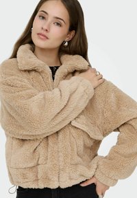 Veste en fausse fourrure beige avec un col montant, une fermeture éclair et une coupe oversize. Présente un tissu texturé et des manches amples.