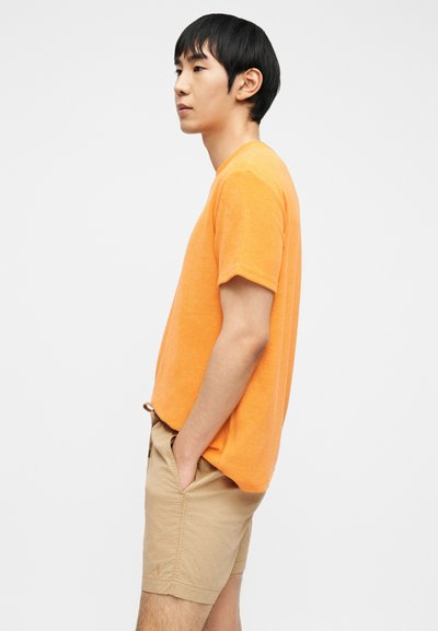 Νεαρός άνδρας με πορτοκαλί κοντομάνικο πουκάμισο και μπεζ σορτς στέκεται πλαγίως με τα χέρια στις τσέπες, μπροστά σε απλό λευκό φόντο.