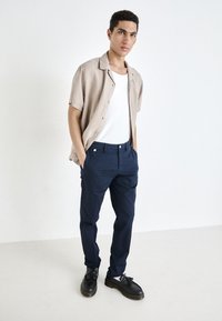 Light beige short-sleeve shirt, white tank top, navy pants, and black shoes with accent details. Smooth fabric, relaxed fit, and tailored design.