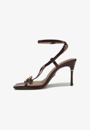 Tamsiai bordo spalvos aukštakulnė sandalų pora su kulkšnies dirželiu, kvadratine nosimi ir dekoratyviniu auksiniu segtuku priekyje ant dirželio.