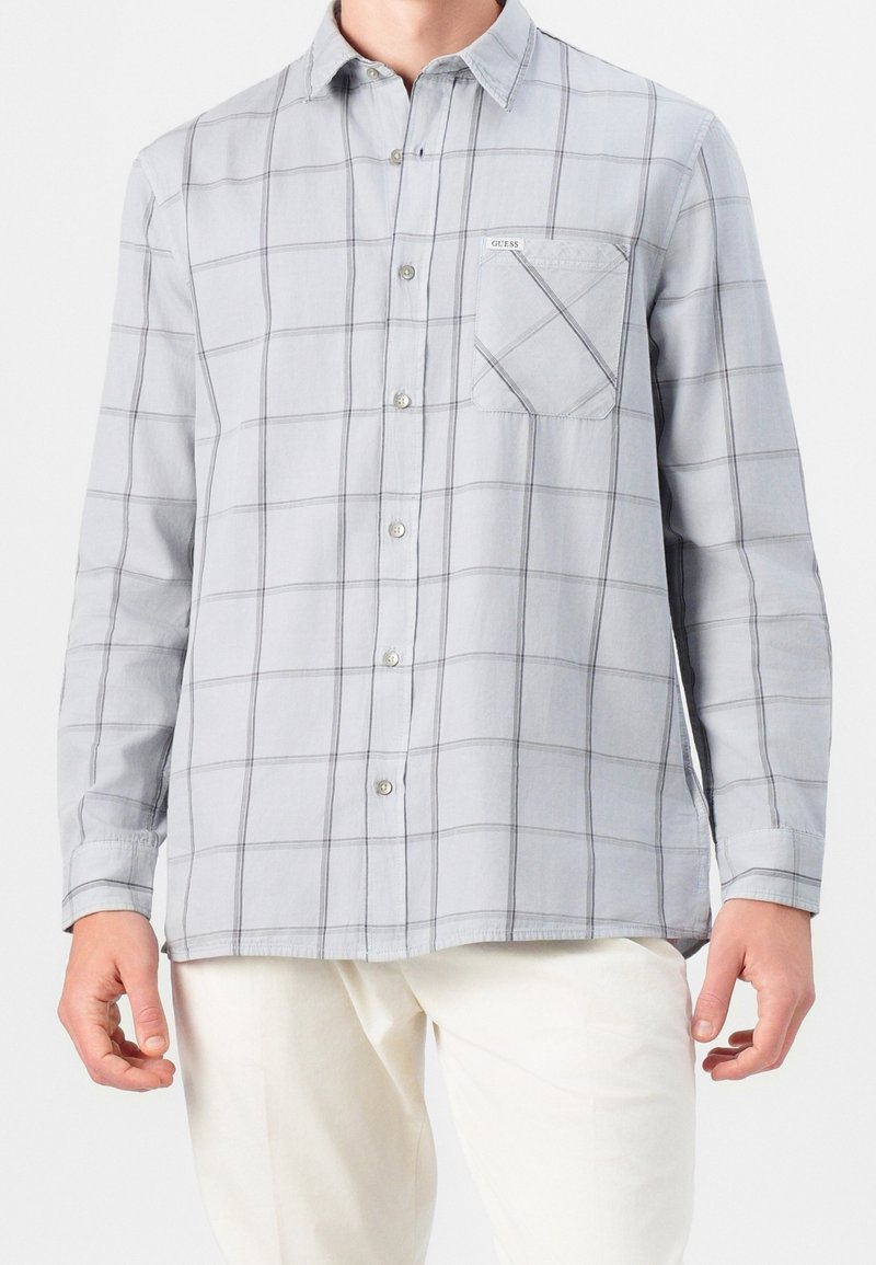 Homme portant une chemise à manches longues gris clair avec un motif à carreaux et une poche avant, associée à un pantalon blanc.