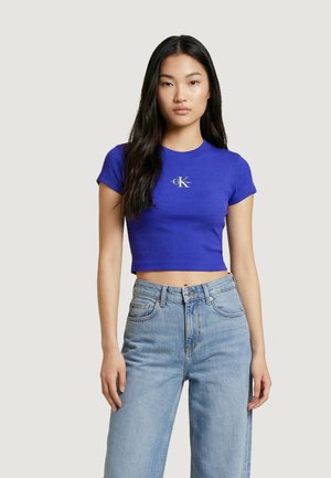 Mladá žena s dlouhými tmavými vlasy nosící přiléhavé modré krátké tričko s logem a světle modré džíny s vysokým pasem, stojící čelem k nám.