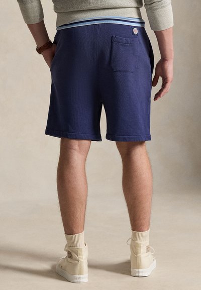 Σκούρο μπλε σορτς με διπλωμένο τελείωμα, ελαστική μέση με μπλε ρίγες και πίσω τσέπη, εμφανιζόμενα από πίσω με ανοιχτό φόντο.