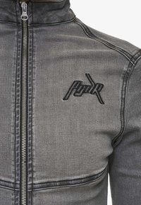 Gray denim jacket with a zip front, high collar, and embroidered logo on the chest. Features visible stitching and a textured finish.
