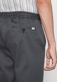 Šedé bavlněné kalhoty s elastickým pasem, zadní kapsou s knoflíkovým uzavřením a malou nášivkou viditelnou na kapse.