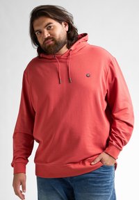 Felpa rosso corallo con cappuccio e lacci, tasca a marsupio e polsini a coste, realizzata in un tessuto morbido e liscio, abbinata a jeans blu.