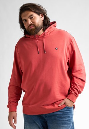 Koraalrode hoodie met trekkoorden, kangaroozak en geribbelde manchetten, gemaakt van een zachte, gladde stof, gecombineerd met blauwe jeans.